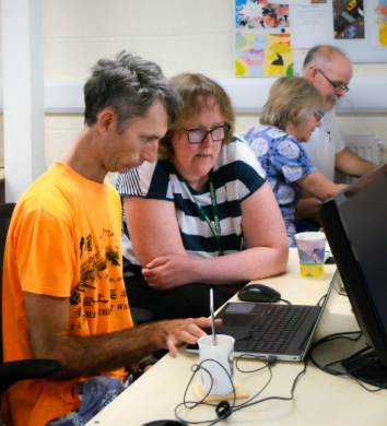 Volunteer Kate sat with a Compaid digital skills client in an orange tshirt at a table. Both are looking at a laptop, while Kate guides the client on using it.